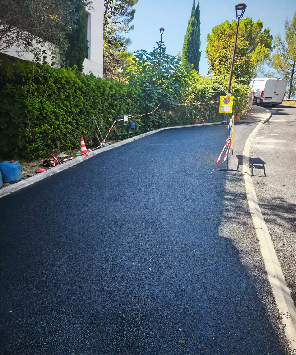 Une voie d'acces neuve en enrobe noir, bordee d'une haie, avec balisage de chantier
